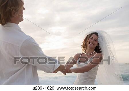 Stock Photograph - Bride and groom on beach, holding hands, fooling around, laughing. Fotosearch