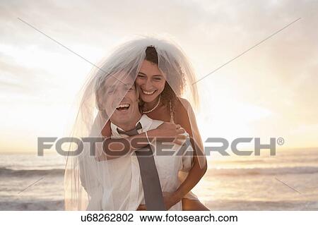 Stock Image - Bride riding piggyback on groom on beach against sunset. Fotosearch