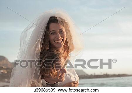 Stock Photo - Bride riding piggyback on groom on beach against sunset. Fotosearch