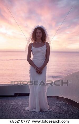 Stock Photo - Bride standing on beach against sunset. Fotosearch