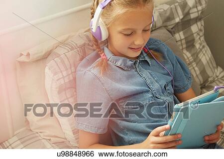 Cropped view of girl sitting on bed wearing headphones looking down at digital tablet smiling View Large Photo Image Stock Photograph - Cropped view of girl sitting on bed wearing headphones looking down at digital tablet smiling. Fotosearch
