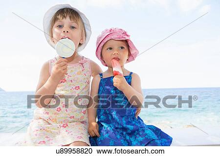 Female toddler and sister wearing sunhats eating ice lollies on beach View Large Photo Image Stock Image - Female toddler and sister wearing sunhats eating ice lollies on beach. Fotosearch