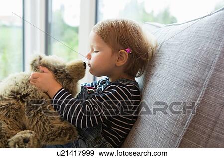 Stock Photo - Female toddler sitting on sofa kissing teddy bear. Fotosearch