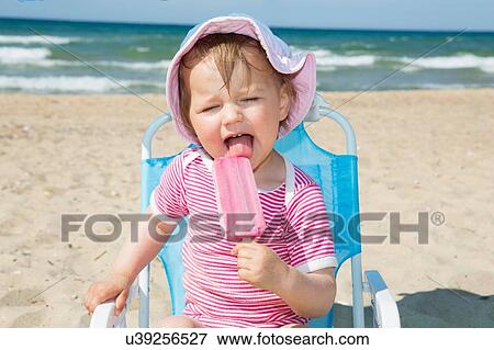 Stock Photo - Female toddler wearing sunhat eating ice lolly on beach chair. Fotosearch