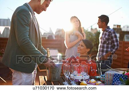 Stock Image - Friends at party on roof terrace preparing champagne. Fotosearch