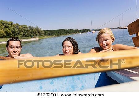 Stock Image - Friends with wet hair in lake holding onto boat looking at camera, Schondorf, Ammersee, Bavaria, Germany. Fotosearch