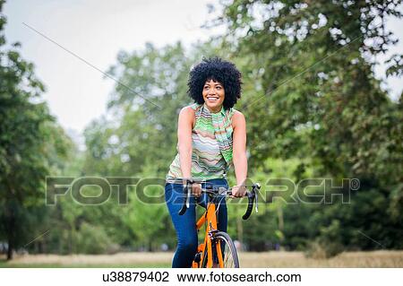 Stock Image - Front view of mature woman with afro riding bicycle looking away smiling. Fotosearch