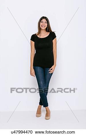 Stock Image - Full length front view of young woman standing legs crossed looking at camera smiling. Fotosearch
