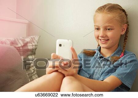 Girl sitting leaning against wall looking at smartphone smiling View Large Photo Image Stock Image - Girl sitting leaning against wall looking at smartphone smiling. Fotosearch