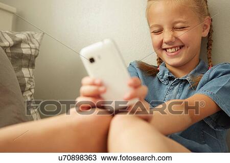Stock Image - Girl sitting leaning against wall holding smartphone, eyes closed smiling. Fotosearch