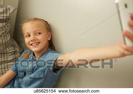 Girl sitting leaning against wall using smartphone to take selfie smiling View Large Photo Image Picture - Girl sitting leaning against wall using smartphone to take selfie smiling. Fotosearch