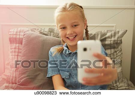 Stock Image - Girl sitting on bed using smartphone to take selfie smiling. Fotosearch