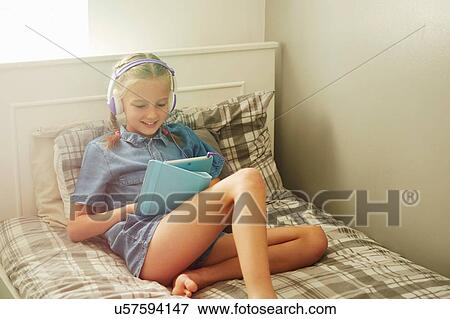 Girl sitting on bed wearing headphones looking down at digital tablet smiling View Large Photo Image Stock Photo - Girl sitting on bed wearing headphones looking down at digital tablet smiling. Fotosearch