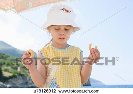 Stock Image - Girl wearing sunhat eating doughnut on beach. Fotosearch