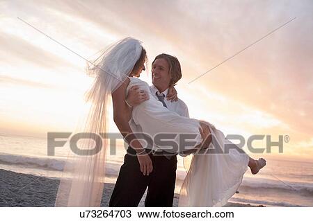 Stock Image - Groom carrying bride on beach against sunset. Fotosearch