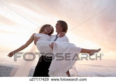 Groom carrying bride on beach against sunset View Large Photo Image Stock Photo - Groom carrying bride on beach against sunset. Fotosearch