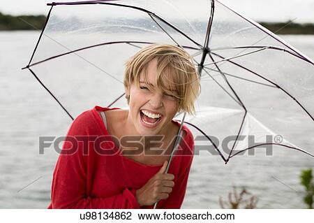 Stock Image - Happy young woman holding transparent umbrella at lake. Fotosearch
