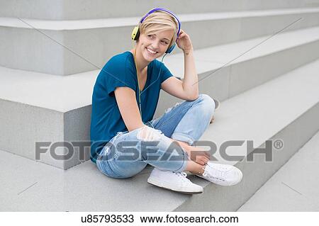 Stock Image - Happy young woman wearing headphones sitting on city stairway. Fotosearch