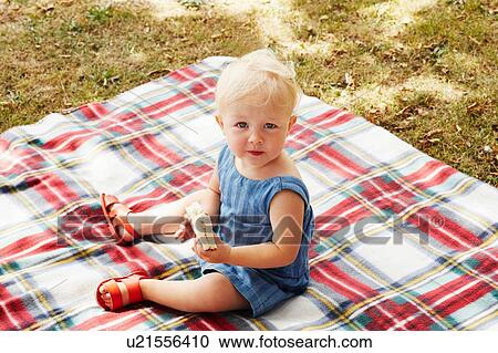 Stock Image - High angle view of baby girl sitting on picnic blanket looking at camera. Fotosearch