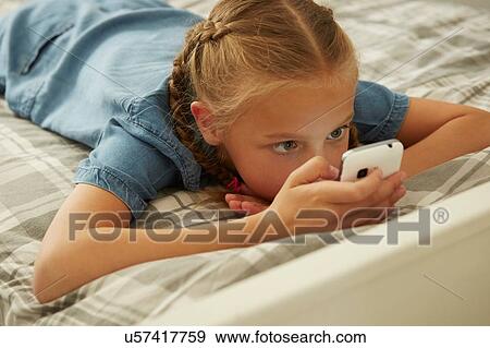 High angle view of girl lying on bed looking at smartphone View Large Photo Image Stock Photo - High angle view of girl lying on bed looking at smartphone. Fotosearch