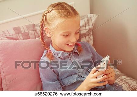 Stock Photograph - High angle view of girl sitting on bed looking down at smartphone smiling. Fotosearch
