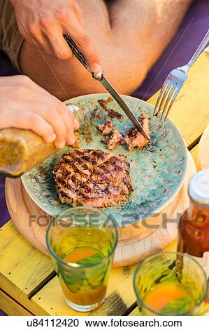 Stock Image - High angle view of two people cutting meat and pouring condiment onto plate. Fotosearch