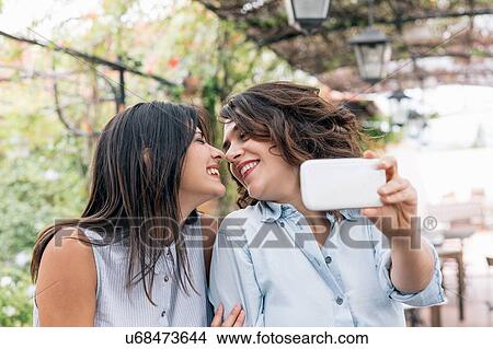 Picture - Lesbian couple in plant covered archway using smartphone to take selfie, face to face, Florence, Tuscany, Italy. Fotosearch