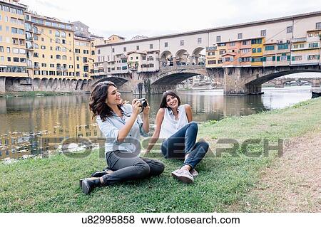 Stock Photo - Lesbian couple sitting on arno river bank in front of Ponte Vecchio holding digital camera smiling, Florence, Tuscany, Italy. Fotosearch