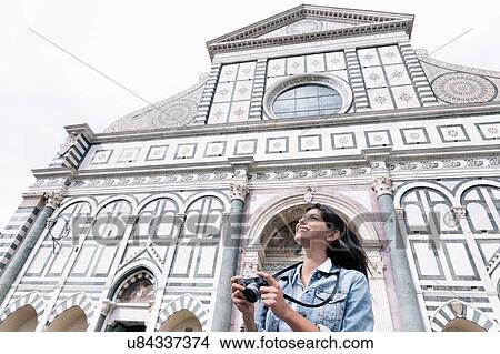 Picture - Low angle view of young woman using digital camera in front of church looking up, Piazza Santa Maria Novella, Florence, Tuscany, Italy. Fotosearch