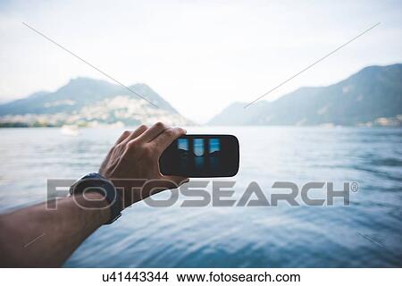 Picture - Male hand photographing with smartphone, Lake Lugano, Switzerland. Fotosearch