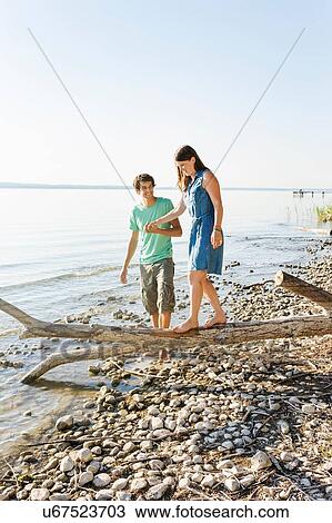 Stock Image - Man helping woman to balance on driftwood, holding hands, Schondorf, Ammersee, Bavaria, Germany. Fotosearch