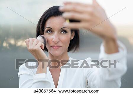 Picture - Mature businesswoman applying blusher using smartphone. Fotosearch