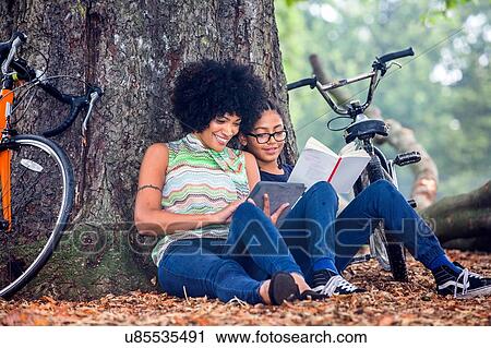 Mature woman and son sitting against park tree reading book and digital tablet View Large Photo Image Stock Image - Mature woman and son sitting against park tree reading book and digital tablet. Fotosearch