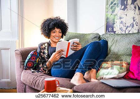 Stock Image - Mature woman relaxing on living room sofa reading book. Fotosearch