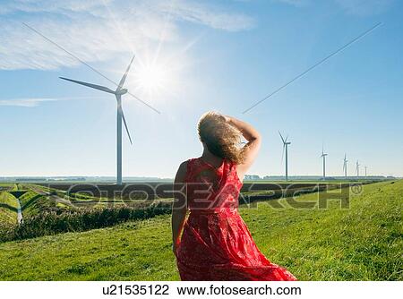 Stock Image - Mature woman standing in field, watching wind turbines on windfarm, rear view, Rilland, Zeeland, Netherlands. Fotosearch