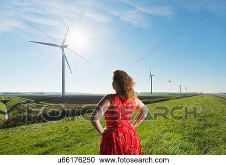 Stock Image - Mature woman standing in field, watching wind turbines on windfarm, rear view, Rilland, Zeeland, Netherlands. Fotosearch