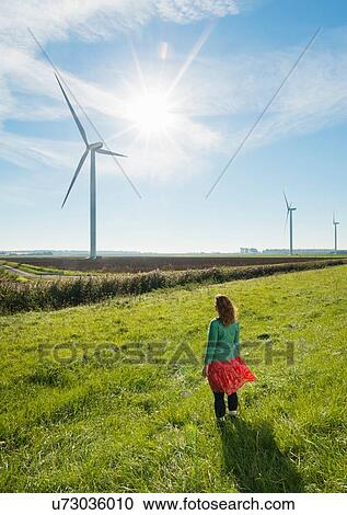 Stock Image - Mature woman standing in field, watching wind turbines on windfarm, rear view, Rilland, Zeeland, Netherlands. Fotosearch