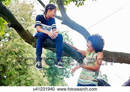 Stock Image - Mature woman watching son sitting on park tree branch. Fotosearch
