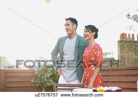 Stock Photo - Mid adult couple standing on roof terrace looking away smiling, mouth open. Fotosearch