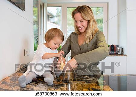 Stock Image - Mid adult woman and toddler daughter preparing dough in kitchen. Fotosearch