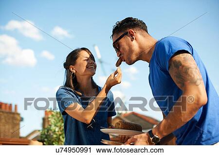 Mid adult woman feeding sausage to boyfriend at rooftop party View Large Photo Image Stock Image - Mid adult woman feeding sausage to boyfriend at rooftop party. Fotosearch