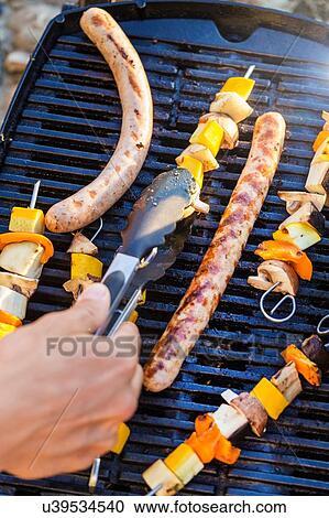 Stock Image - Person using tongs to turn sausages and kekabs on barbecue griddle. Fotosearch