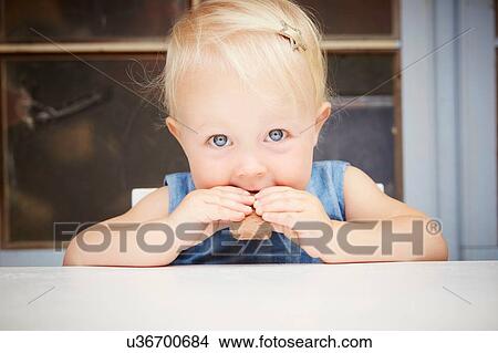 Picture - Portrait of cute blonde baby girl looking at camera eating chocolate biscuit. Fotosearch