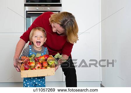 Stock Photo - Portrait of female toddler and mother holding crate of apples in kitchen. Fotosearch