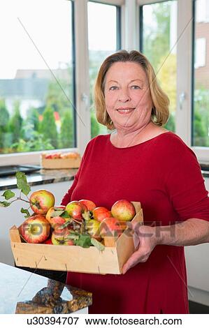 Portrait of senior woman holding crate of apples in kitchen View Large Photo Image Stock Photo - Portrait of senior woman holding crate of apples in kitchen. Fotosearch