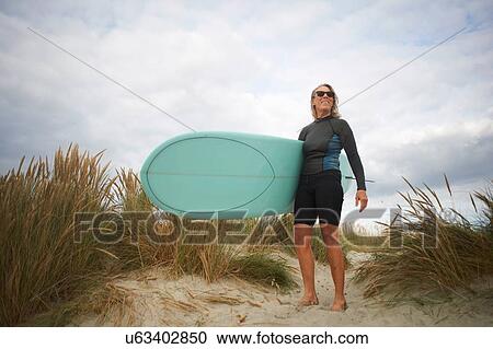 Stock Image - Portrait of senior woman on sand, holding surfboard. Fotosearch