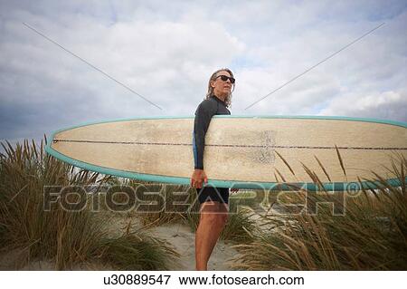 Stock Photo - Portrait of senior woman on sand, holding surfboard. Fotosearch