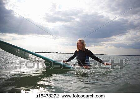 Stock Image - Portrait of senior woman sitting on surfboard in sea, smiling. Fotosearch