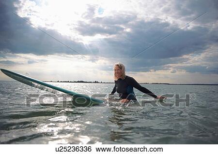 Stock Photograph - Portrait of senior woman sitting on surfboard in sea, smiling. Fotosearch