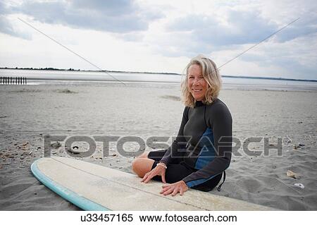 Stock Photography - Portrait of senior woman sitting on beach next to surfboard. Fotosearch